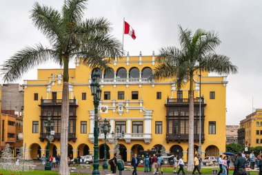 Lima, Peru - 4 Aralık 2008: Plaza de Armas 'ın yeşil çimlerinin ve palmiye ağacının arkasındaki Hükümet Sarayı cephesi, gümüş gökyüzü altında üst düzey kırmızı-beyaz Peru bayrağıyla sarıdır. Yaya arabaları ön tarafta..
