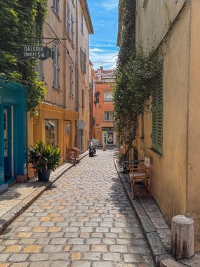 Saint Tropez, France - July 13, 2024: Downtown scenery. Rue du Cloche with small businesses one being Gallerie Henri Sie. 4-story residenial beuildings form narrow canyon. Green foliage