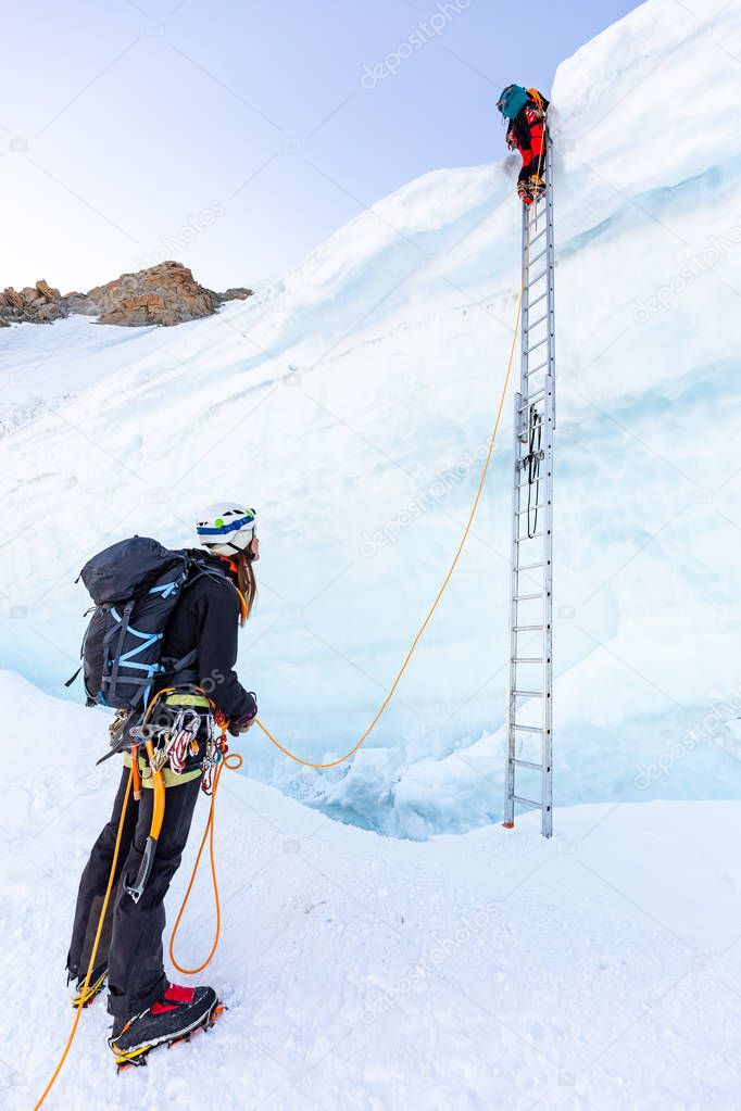 Two alpinists mountaineers climb ladder over ice crevasse.