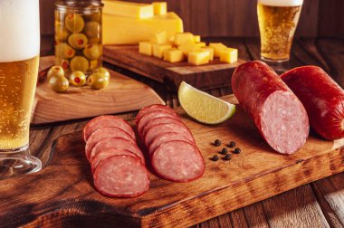 Kesme tahtasına dilimlenmiş füme salam. İki bardak bira, bir avuç peynir ve zeytin.