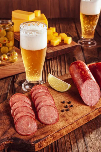 Kesme tahtasına dilimlenmiş füme salam. İki bardak bira, bir avuç peynir ve zeytin.