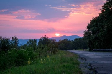 Güneş, dağların arka planında doğar. Uzağa uzanan yeşil çimenler ve bir yol. Stanitsa Dakhovskaya, Aygea Cumhuriyeti, Rusya.
