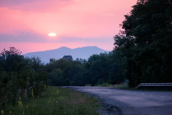 the sun rises against the background of the mountains. Green grass and a road stretching into the distance. Stanitsa Dakhovskaya, Republic of Adygea, Russia.