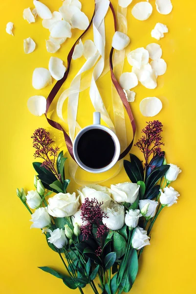 Cup of coffee on the bright yellow background with flower bouquet, petals and tapes. Breakfast on Birthday, Womans or Mothers Day. Flat lay styling