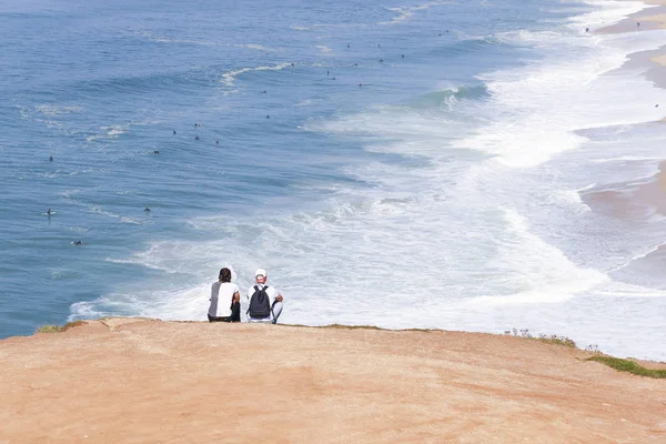 Two people sit near the ocean and look at the surfers.