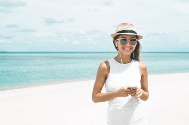 Güzel körfezde arkadaşlarla iletişim kurmak ve müzik dinlemek için telefon ve kulaklık kullanarak yürüyen genç bir kadın.