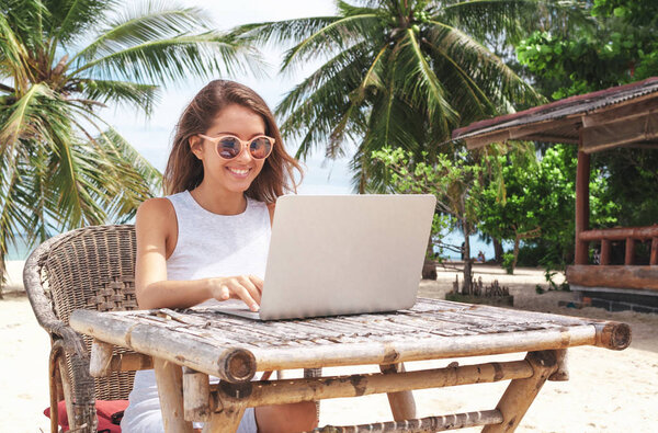 Молодая женщина, работающая с ноутбуком на тропическом пляже острова под пальмами
