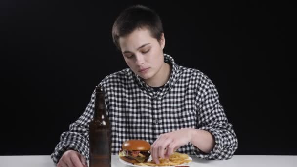 Chica joven inusual está comiendo papas fritas, hamburguesa en el plato, botella de cerveza en la mesa, concepción de la dieta, fondo negro — Vídeos de Stock