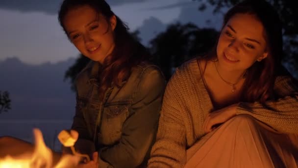 Dos jóvenes caucásicas se sientan por la noche junto al fuego en la naturaleza, cocinando marshmelow, sonriendo, comiendo — Vídeos de Stock