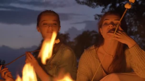 Twee jonge Kaukasische meisjes zitten 's avonds bij het haardvuur in de natuur, eten van marshmelow, een open haard te kijken, denken — Stockvideo