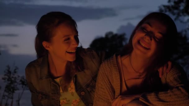Duas jovens caucasianas sentam-se à noite ao lado do fogo na natureza, rindo, sorrindo — Vídeo de Stock