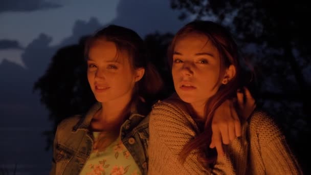 Duas jovens caucasianas sentam-se à noite perto do fogo na natureza, olham para a câmera, retrato — Vídeo de Stock