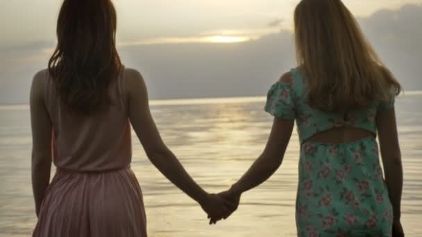 Zwei junge kaukasische Mädchen in Kleidern, die bei Sonnenuntergang am seichten Wasser entlanggehen, sich umdrehen und in die Kamera schauen — Stockvideo