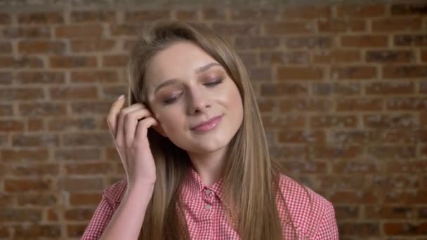 Menina confiante jovem toca seu cabelo, assistindo a câmera, fundo de tijolo — Vídeo de Stock