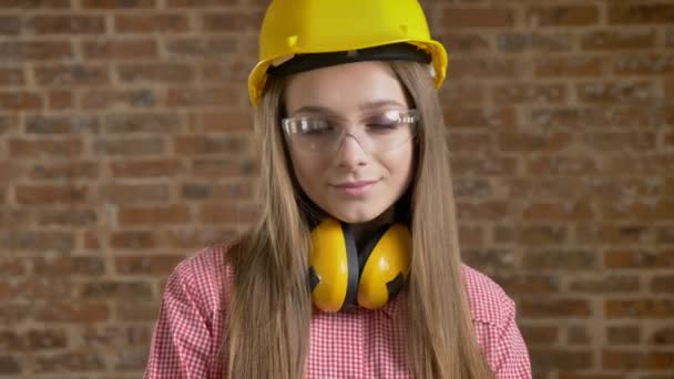 Young attractive girl builder is nodding her head and glances away, watching at camera, brick background — Stock Video