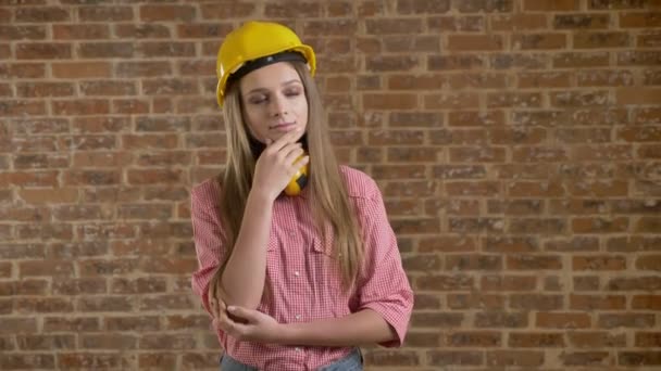 Young beautiful girl builder standing and evaluating something, thinking process, brick background — Stock Video