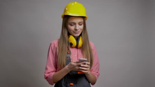 Jovem construtor menina bonita está mostrando tela croma de seu smartphone, sorrindo, conceito de comunicação, fundo cinza — Vídeo de Stock