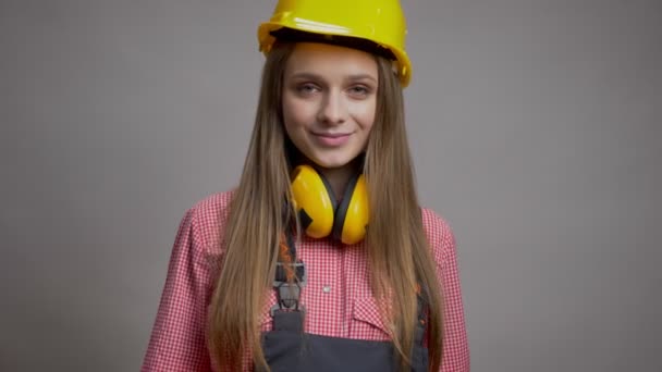 Молодая улыбающаяся девушка строитель показывает большой палец вверх, как знак, концепция общения, серый фон — стоковое видео