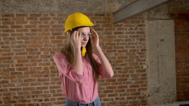 Jovem slim girl builder está vestindo glases, movendo as mãos através do corpo, confiança, assistindo a câmera, fundo de tijolo — Vídeo de Stock