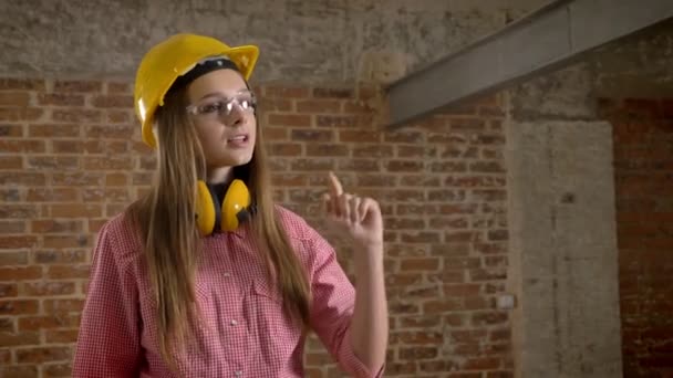 Joven constructor chica confiada apunta al trabajo y muestra el pulgar hacia arriba, como signo, fondo de ladrillo — Vídeos de Stock
