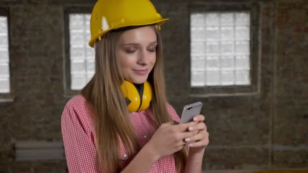 Joven hermosa niña sonriente constructor está utilizando su teléfono inteligente, watchig en la cámara, concepto de comunicación — Vídeos de Stock