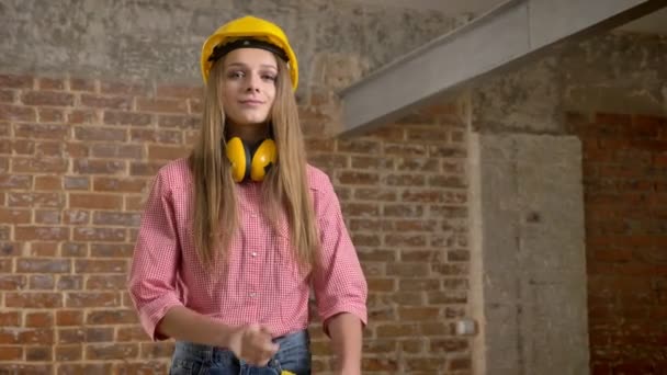 Jong mooi meisje bouwer overschrijdt wapens, lacht, kijken op camera, baksteen achtergrond — Stockvideo