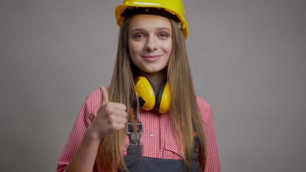 Jovem menina feliz construtor mostra polegar para cima, como sinal, rindo, conceito de comunicação, fundo cinza — Vídeo de Stock