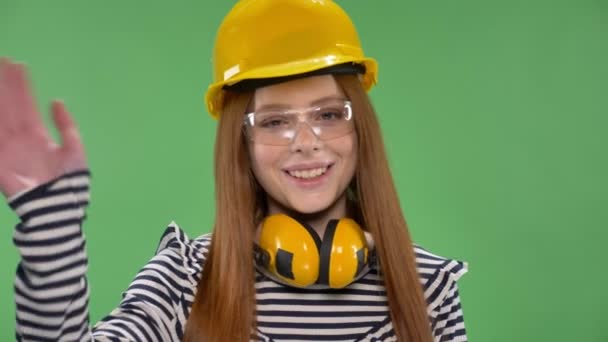 Молодая улыбающаяся рыжая девушка в строительном шлеме, наушниках, очках, машет рукой, знак "привет" — стоковое видео