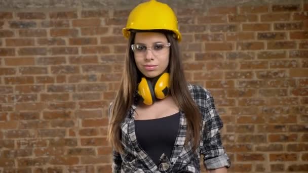 Молодые довольно привлекательные женщины в шлеме смотрят прямо в камеру и снимают очки, женщина-строитель, кирпичный фон — стоковое видео