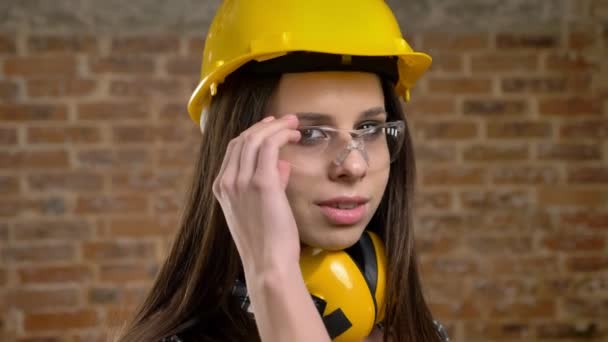 Jóvenes mujeres bastante atractivas en casco y auriculares mirando directamente a la cámara y sosteniendo sus gafas, constructora femenina, fondo de ladrillo, primer plano — Vídeo de stock