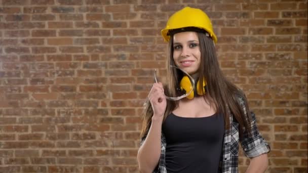 Hermosas mujeres jóvenes atractivas en casco y auriculares mirando directamente a la cámara y sosteniendo sus gafas, sonriendo, constructora femenina, fondo de ladrillo — Vídeo de stock