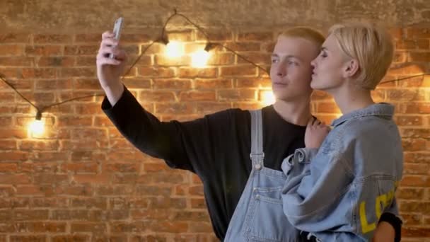 Jovem casal atraente de hipsters faz selfie no smartphone, concepção de comunicação, fundo de tijolo — Vídeo de Stock