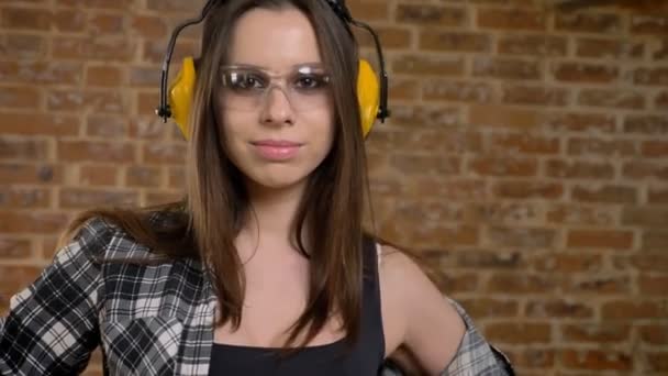 Hermosas mujeres jóvenes atractivas en gafas, auriculares, posando frente a la cámara, dando la vuelta, constructora femenina, fondo de ladrillo — Vídeo de stock