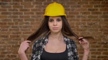 Genç güzel doğruca kameraya bakarak ve dokunaklı onun uzun saç, kadın builder, tuğla arka plan kask içinde çekici kadınlar