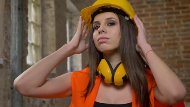 Mujeres jóvenes bastante atractivas en casco y auriculares mirando en la cámara, tocando y ajustando la casque, cruzando las manos, mujer constructora, ladrillo edificio fondo — Vídeos de Stock