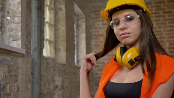 Junge hübsche Frauen mit Helm, Kopfhörer und Brille, die in die Kamera blicken, ihre langen Haare berühren, lächeln, Bauarbeiterin, Backsteinbau Hintergrund — Stockvideo
