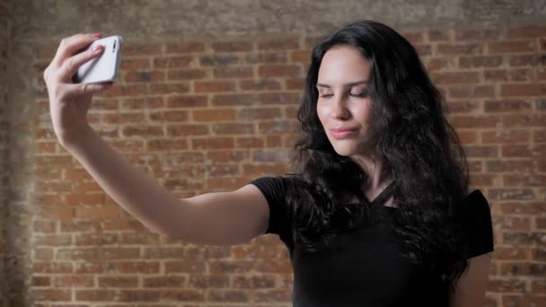 Junge schöne brünette Mädchen macht Selfie auf ihrem Smartphone, lächelt, Kommunikationskonzeption, Seitenansicht, Backstein Hintergrund — Stockvideo