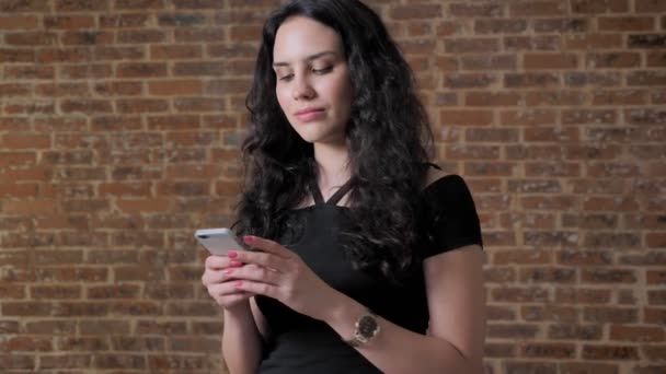 Joven hermosa chica morena está enviando mensajes de texto en su teléfono inteligente, concepto de comunicación, ladrillo backgroung — Vídeos de Stock