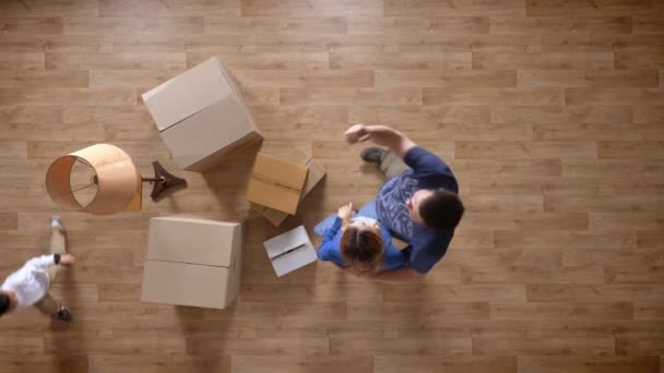 Jovem família de pé em casa com caixas, marido apontando e mostrando algo para a esposa, criança correndo em torno de topo tiro parquet piso de madeira — Vídeo de Stock