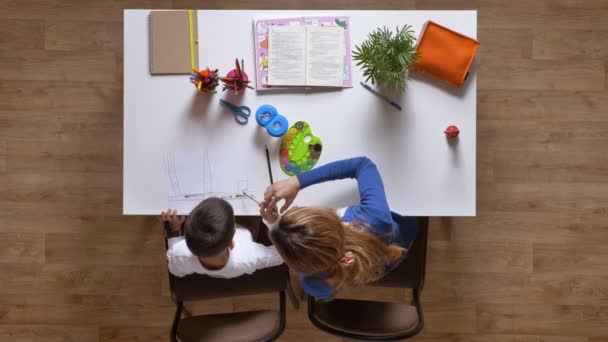 Joven madre pintando con su hijo, sentado detrás de la mesa, niño haciendo tareas de parquet plano piso de madera — Vídeos de Stock