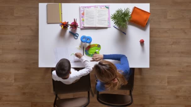 Jovem mãe pintura com filho, explicando a criança, sentado atrás da mesa, menino fazendo lição de casa top shot parquet piso de madeira — Vídeo de Stock