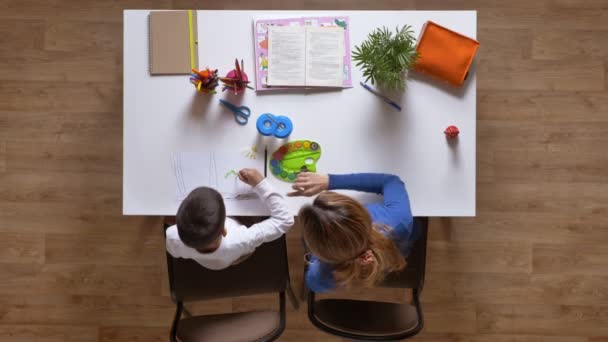 Jonge moeder schilderen met zoon, uit te leggen om de kid, zittend achter tafel, jongen doen van huiswerk top neergeschoten houten parketvloer — Stockvideo