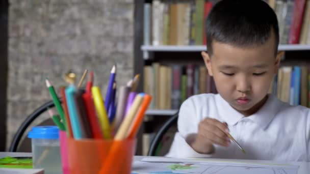 Маленький азиатский мальчик рисует на бумаге и сидит за столом, ребенок делает домашнее задание, книжные полки фон — стоковое видео