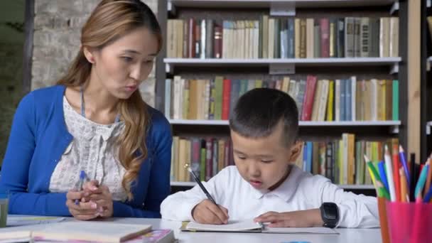 Молодая мать помогает сыну в учебе, женщина разговаривает с ребенком, сидит за столом, азиатский ребенок делает домашнее задание, книжные полки фон — стоковое видео