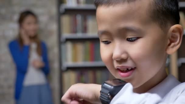 Pouco asiático menino falando com a mãe através de relógio inteligente, prateleiras de livros e parede de tijolo fundo — Vídeo de Stock