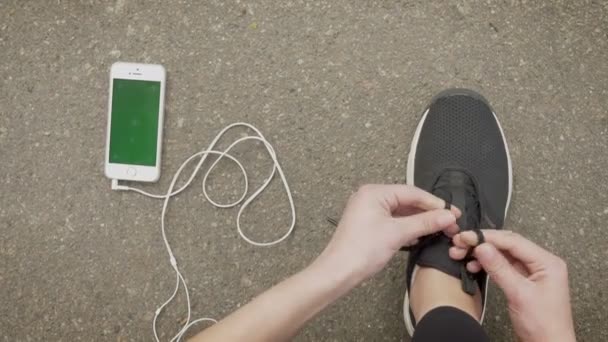 Menina desportista amarra seus atacadores enquanto seu smartphone e fones de ouvido estão deitados no chão, tela chroma, conceito de comunicação — Vídeo de Stock