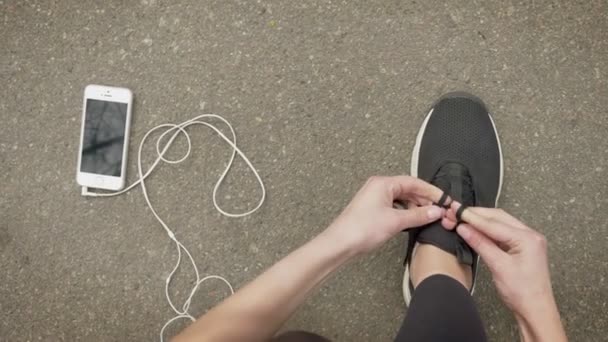 Chica deportista ata sus cordones de zapatos mientras su teléfono inteligente y auriculares están acostados en el suelo, concepto de comunicación — Vídeo de stock