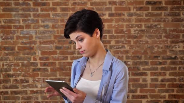 Joven hermosa chica está jugando en su tableta, entretenimiento, concepto de comunicación, fondo de ladrillo — Vídeos de Stock