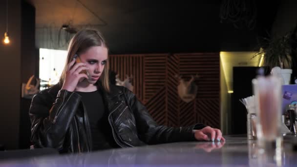 Jonge mooie vrouwen in zwarte jas met zware make-up zitten en praten over telefoon, boos, bar achtergrond — Stockvideo