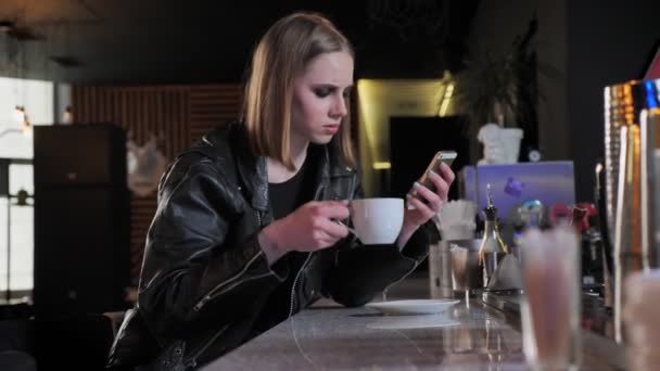 Unga vackra kvinnor i svart jacka med tung make up titta på telefonen och dricka kaffe, bar bakgrund — Stockvideo